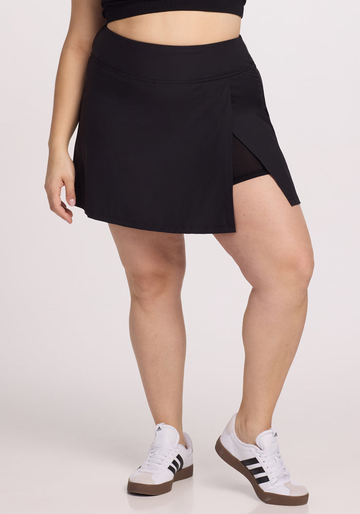 A person is wearing the Woolx Topanga Skort in black, featuring a side slit that reveals black shorts beneath. They pair it with white and black sneakers against a plain, light background. 
