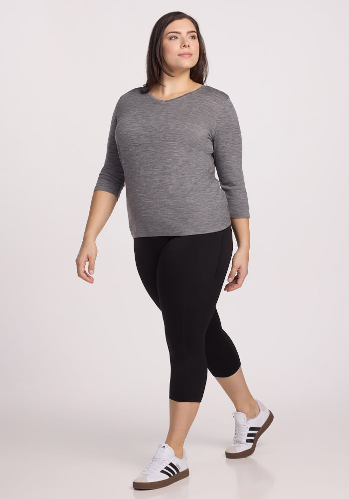 A person in a Woolx Elena 3/4 Sleeve V Neck - Graphite Heather, black leggings, and white sneakers with black stripes walks. They have shoulder-length dark hair and a neutral expression against a plain white background.