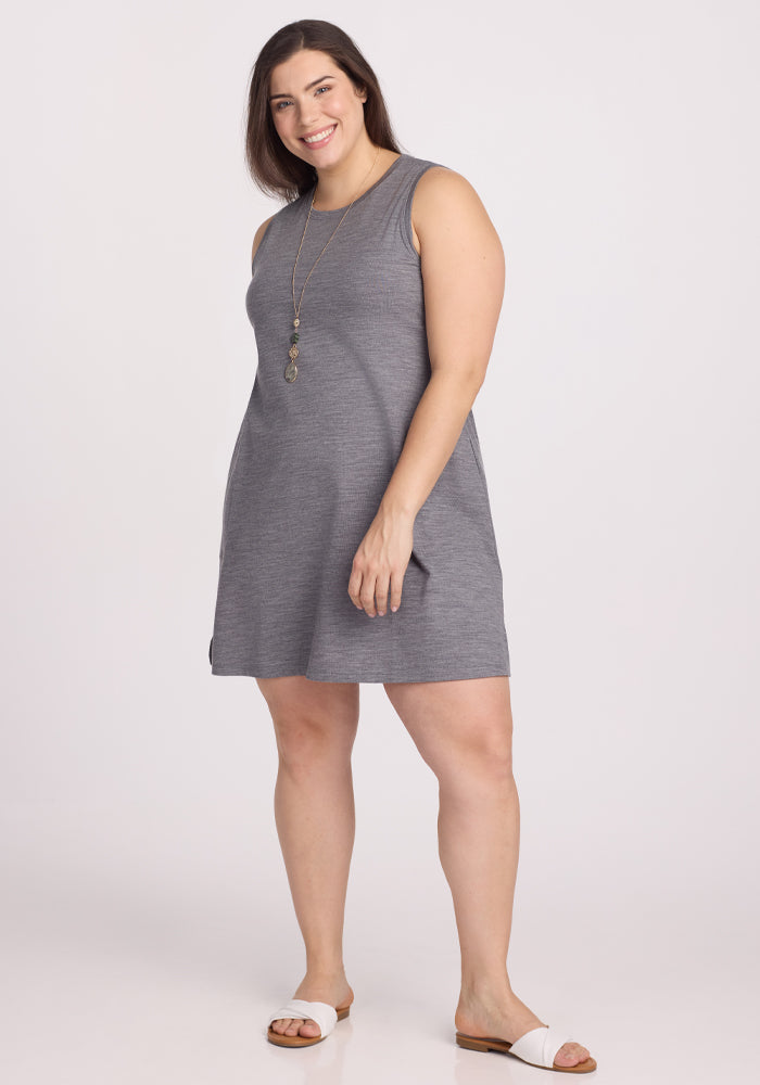 A smiling woman with long hair wears the sleeveless, temperature-regulating Clara Dress in Graphite Heather by Woolx and white sandals. She stands against a plain white background, accessorized with a long necklace. 