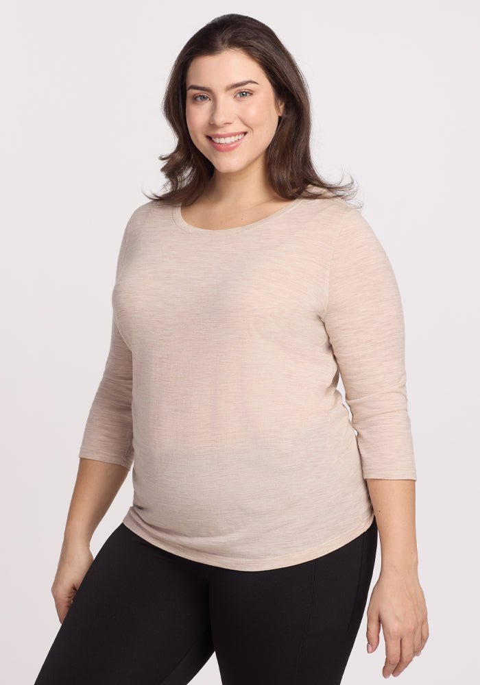 A smiling person with long dark hair stands against a neutral background, wearing Woolx's Jenny 3/4 Sleeve Crew Neck in Cream Heather with black pants. 