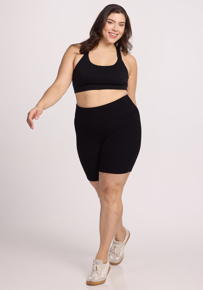 A person is confidently walking while smiling, dressed in Woolx's Miley Bike Shorts - Black and a black sports bra. They have long dark hair, wear white sneakers, and stand against a plain background that highlights their athletic attire.