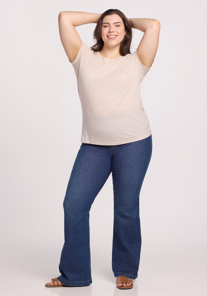 A confident person smiles with hands behind their head, wearing a Woolx Liza Crew Neck Tee in Cream Heather, paired with blue flared jeans and brown sandals against a plain white background.