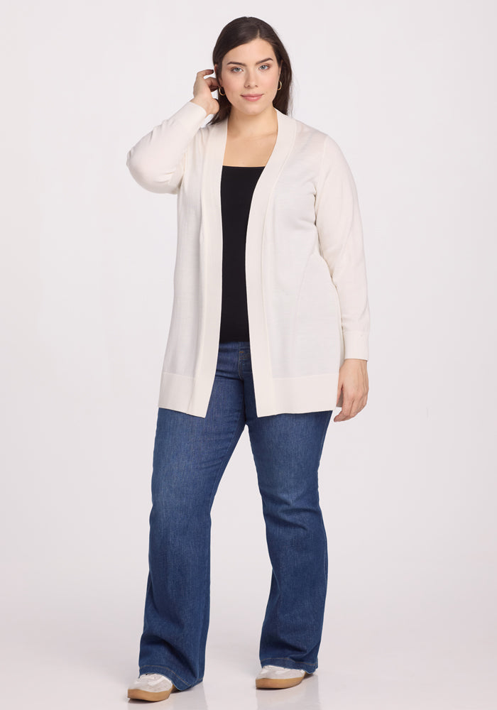 A person wearing the Woolx Macie Sweater in Dewdrop Pearl over a black top and blue jeans stands against a plain white background. They look relaxed, touching their hair with one hand.