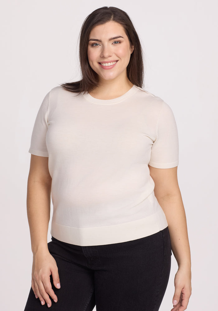A smiling person with long dark hair wears the Woolx Anna Crew Sweater in Dewdrop Pearl, paired with dark pants, standing against a plain light background. 