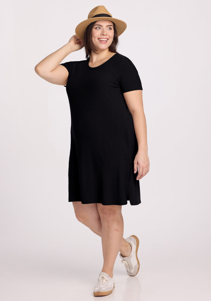 A person confidently poses in the Harlowe Ribbed Dress - Black by Woolx, paired with white sneakers. They smile with one hand on their straw hat against a plain white background, accentuating their casual and stylish look. 