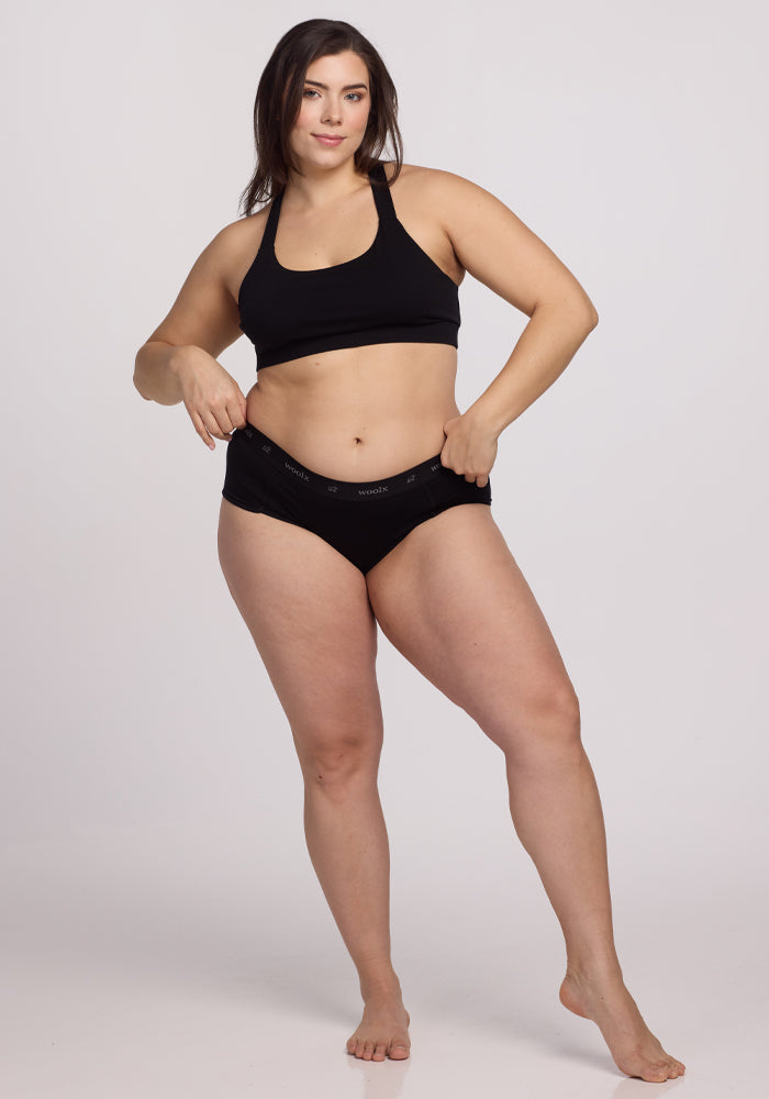 A person stands confidently against a plain background in Woolx's Lila Boy Shorts - Black, featuring lightweight fabric. Barefoot, they embody all-day comfort with one hand on their hip.