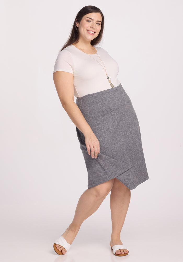 A person smiles at the camera, wearing a white T-shirt and the Woolx Marilyn Flowy Midi Skirt in Graphite Heather. They have long dark hair, accessorized with a long necklace, and stand against a plain white background, completing the look with white sandals.