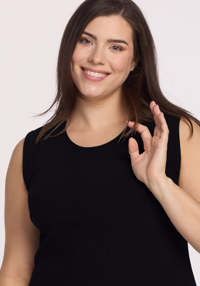 A smiling person with long brown hair wears the Iris Ribbed Dress - Black by Woolx, making an "OK" gesture against a plain background.