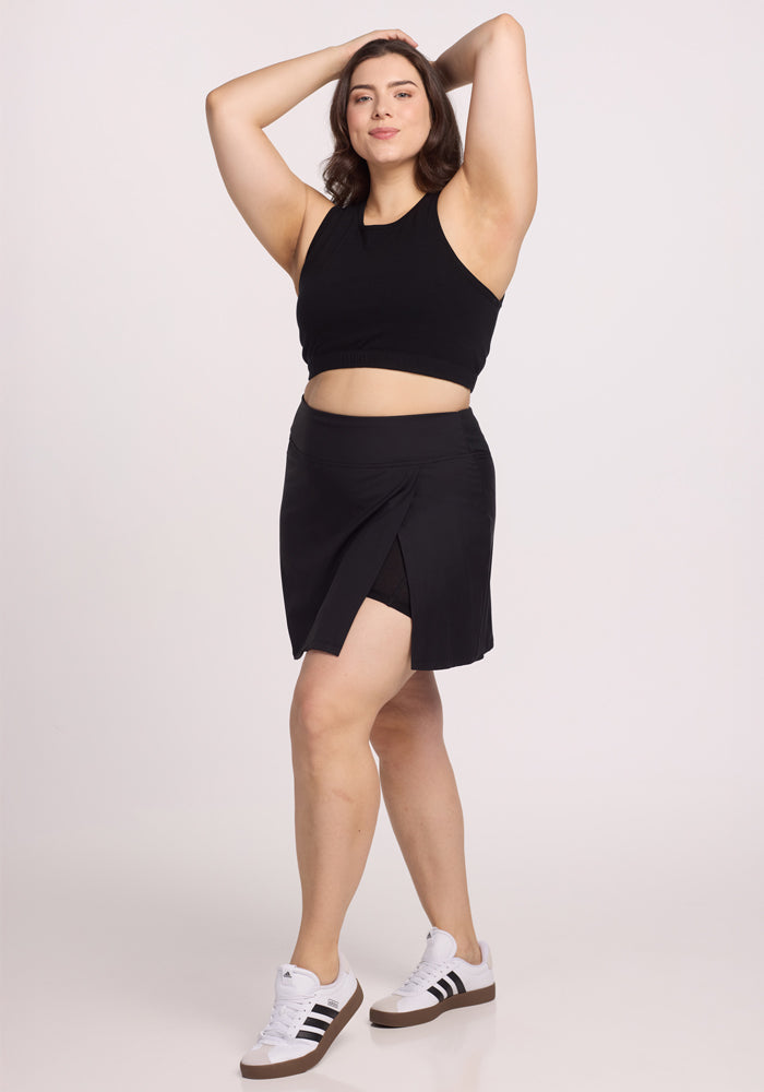 A person confidently poses with hands behind their head, wearing Woolx's black Topanga Skort with a front slit and a sleeveless black top. They have white sneakers featuring black stripes and are smiling against a plain background.