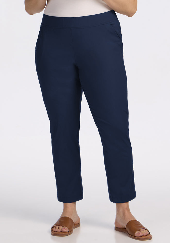 A person wearing Woolx Nicole Tall Ankle Pant in Deep Navy, paired with a white top and brown open-toe sandals, stands against a plain light background.