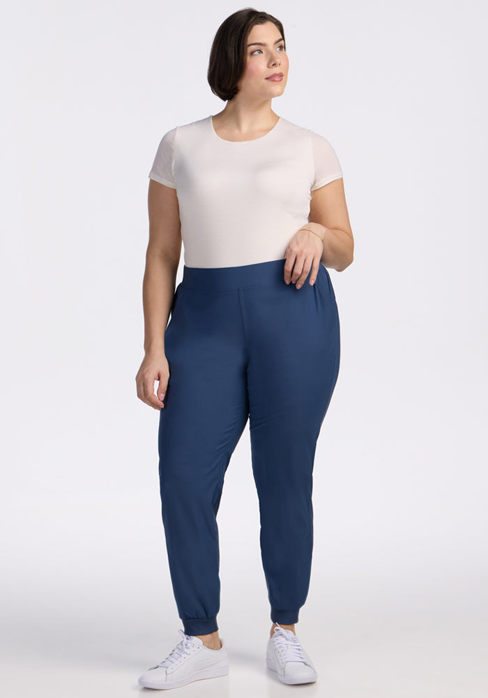 A woman with short brown hair wears a fitted white short-sleeve top, Woolx Palmer Joggers in Vintage Indigo, and white sneakers, standing against a plain white background.