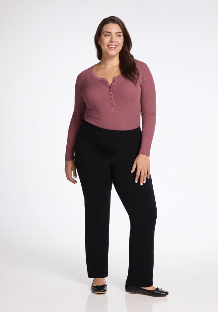 A woman stands smiling, wearing a long-sleeve, maroon top paired with the stylish Woolx Emerson Straight Leg Pants Tall in black. She has long, dark hair and is posing confidently with one hand on her hip while sporting black flats against a white background.