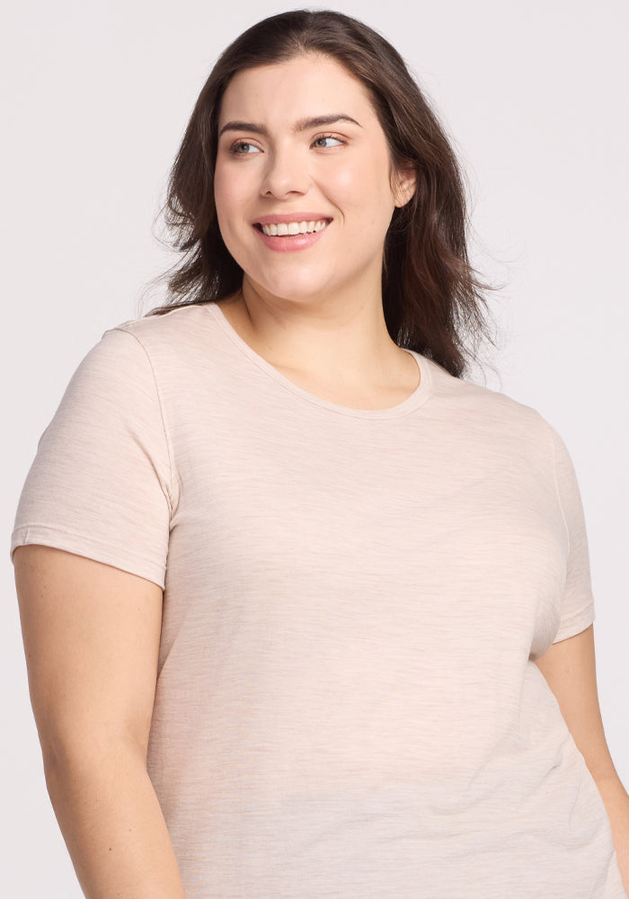 A person with long brown hair smiles and gazes to the side, wearing the Liza Crew Neck Tee in Cream Heather by Woolx. The short-sleeved, lightweight fabric shirt and elegant crew neck stand out against a plain, light-colored background.