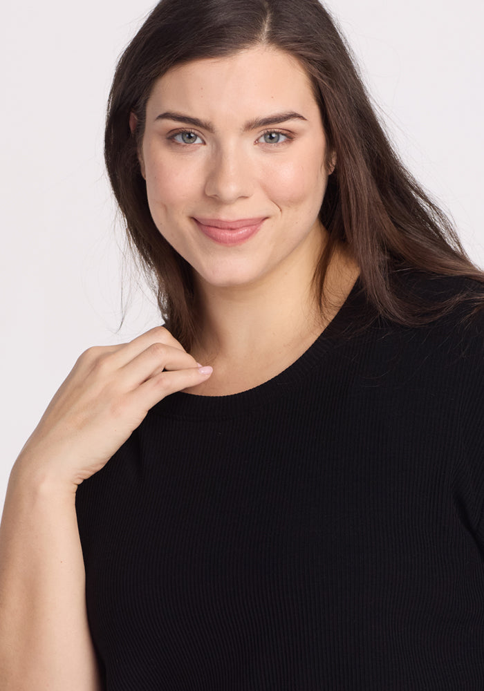 A woman with long brown hair and light skin smiles at the camera, wearing the Woolx Vera Ribbed Tunic in black—a relaxed-fit top. Her left hand rests near her collarbone against a plain, light-colored background.