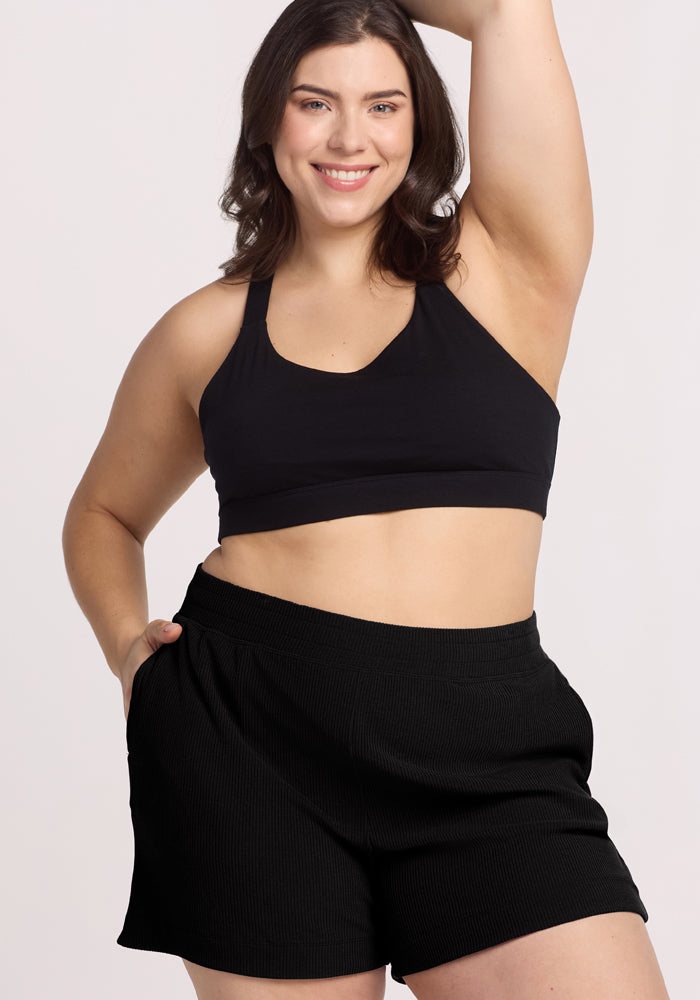 A woman models the Woolx Maisey Ribbed Shorts in black, confidently posing with one hand on her hip and the other raised above her head, smiling against a plain backdrop.