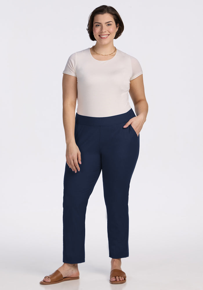 A woman with short brown hair wears a white t-shirt, Woolx Nicole Ankle Pants in Deep Navy with a tapered-leg fit, and brown sandals. She stands smiling with one hand in her pocket against a plain white background.