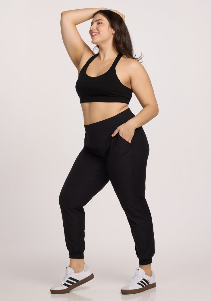 Wearing Woolx's Palmer Joggers - Black, a woman stands confidently against a plain white background, with one hand on her head and the other in the deep pockets of her comfortable joggers. She's smiling, sporting a black sports bra and white sneakers with black stripes.