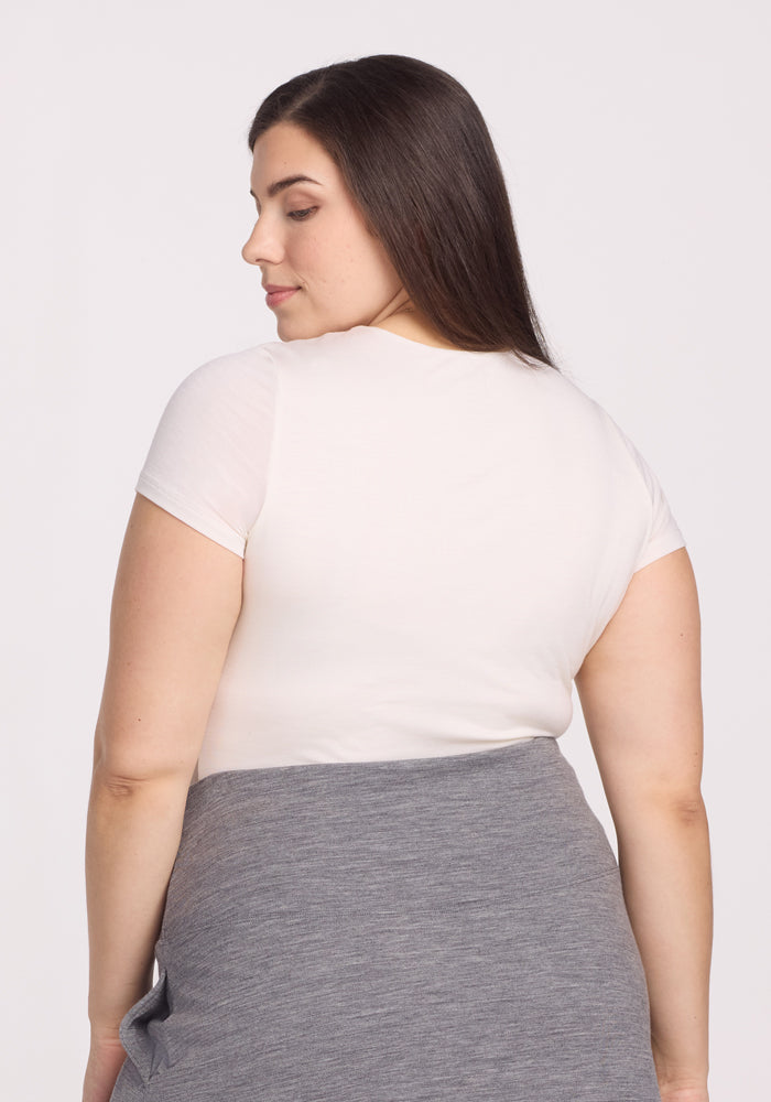 A woman with long, straight brown hair faces away from the camera, wearing the Woolx Gracie Bodysuit in Vanilla for temperature-regulating comfort. A gray garment is tied at her waist. The background is plain and light-colored.