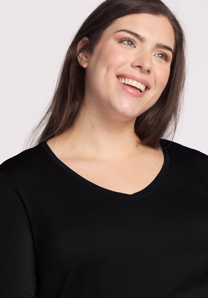A person with long dark hair smiles while looking to the side, wearing the black Woolx Millie Long Sleeve top made of moisture-wicking fabric against a plain, light-colored background.