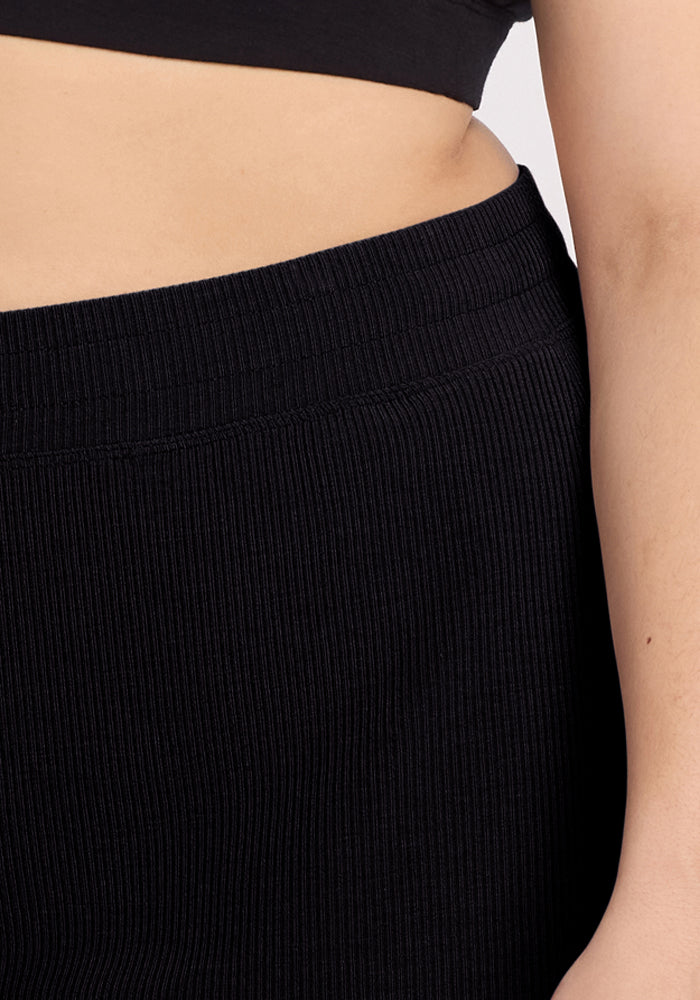 A person is wearing Woolx's Maisey Ribbed Shorts in black with a matching crop top, partially revealing their midriff. The shorts have a visibly ribbed texture against a plain, light-colored background.