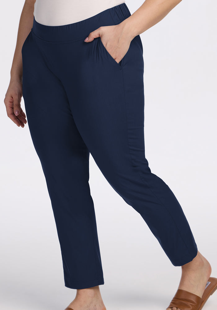 A person wears Woolx Nicole Ankle Pants in Deep Navy with a tapered-leg fit, paired with a white top and tan slide sandals, standing against a plain light background.