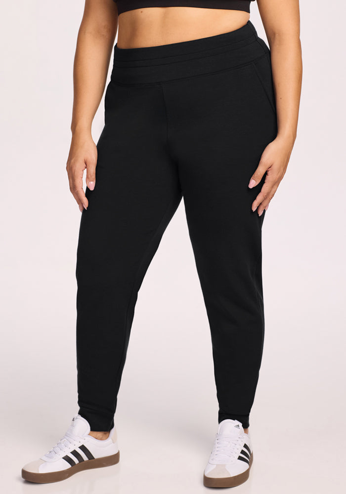 A person wearing Woolx Willa Joggers in black, paired with a black crop top and white sneakers with black stripes, stands against a light background with their hands relaxed by their sides. 