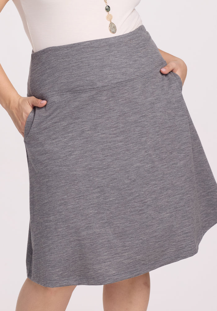 A person wears the Woolx Marilyn Flowy Midi Skirt in Graphite Heather, with hands in pockets. The outfit is styled with a white top and a long beaded necklace against a plain white background.