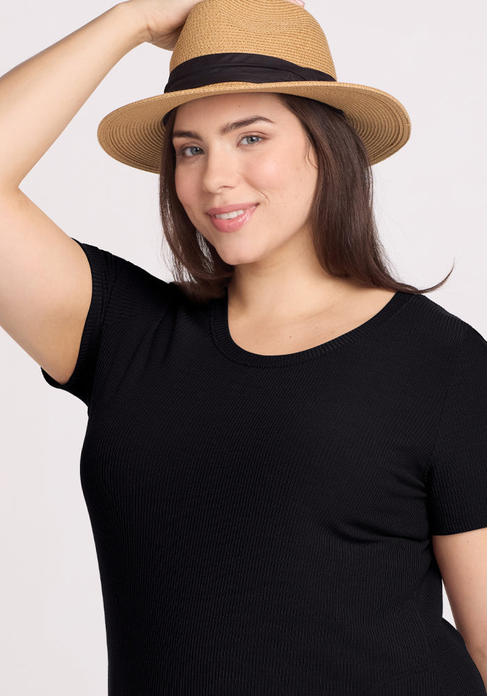 A person wearing the Harlowe Ribbed Dress in black by Woolx gently smiles while adjusting a straw hat with a black band, set against a plain, light-colored background.