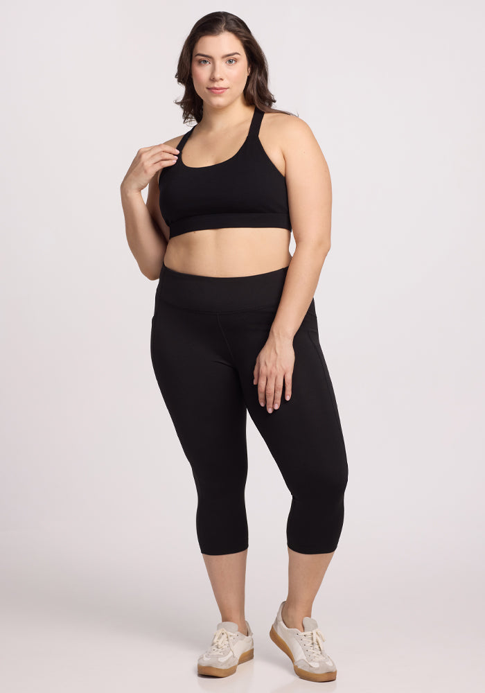 A confident person poses against a plain white background, wearing the Woolx Livi Bra in black. They touch their hair with one hand, placing the other on their waist. Their outfit is completed with odor-resistant leggings and white sneakers featuring beige soles.