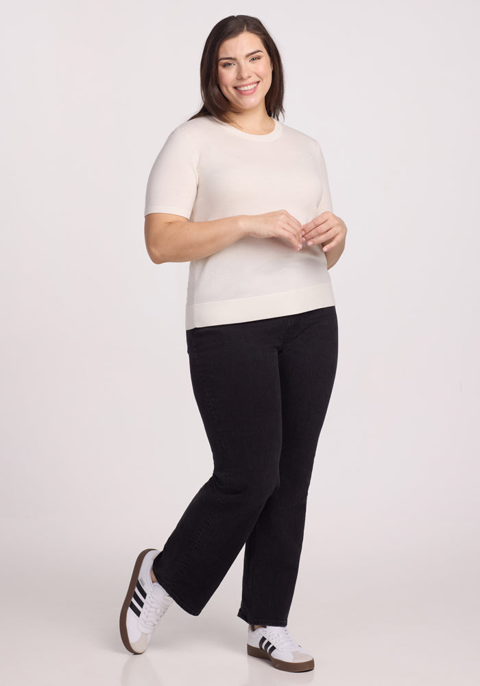 Standing with a smile and one foot slightly forward, the person wears the Woolx Anna Crew Sweater in Dewdrop Pearl, paired with black pants and white sneakers featuring dark stripes against a plain white background.