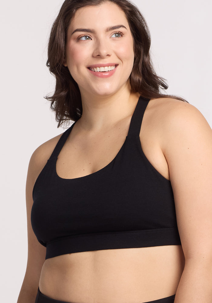 A woman with long dark hair smiles while wearing a Woolx Livi Bra in black, known for its odor resistance. She poses against a plain, light-colored background.