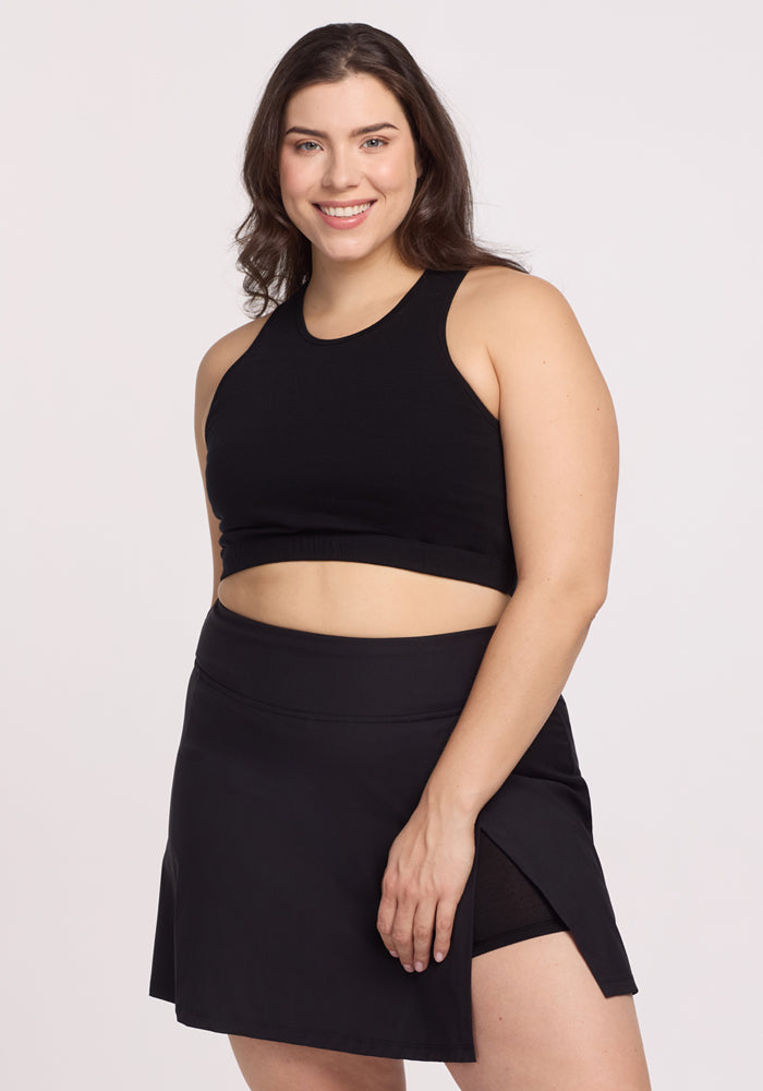 A woman with long, wavy hair stands against a plain white backdrop wearing the Topanga Skort in black by Woolx, smiling in a black sleeveless crop top.