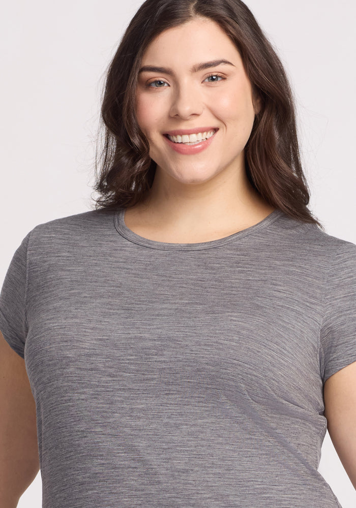 A woman with medium-length brown hair and light skin smiles at the camera, wearing the Woolx Addie Crew Neck in Graphite Heather. The background is plain and light-colored.