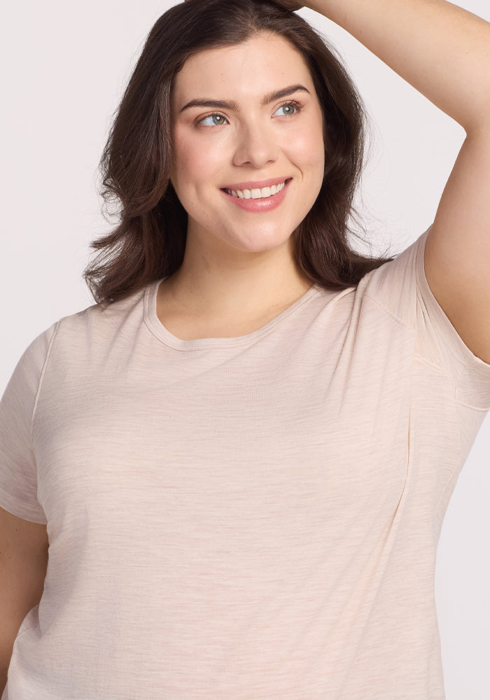 A woman with long brown hair smiles and looks to the side, wearing a Woolx Liza Crew Neck Tee in Cream Heather. She exudes elegance, with one hand raised above her head against a plain, light-colored background.