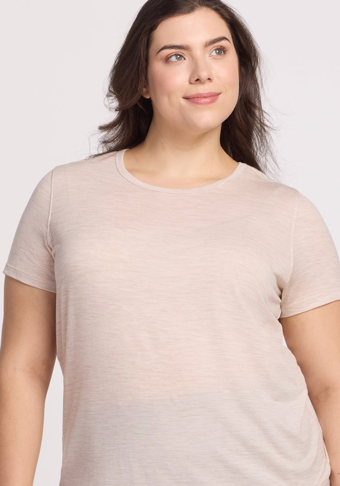 A woman with long dark hair wears the Woolx Liza Crew Neck Tee in Cream Heather, smiling slightly as she looks to the side against a plain, light-colored background.