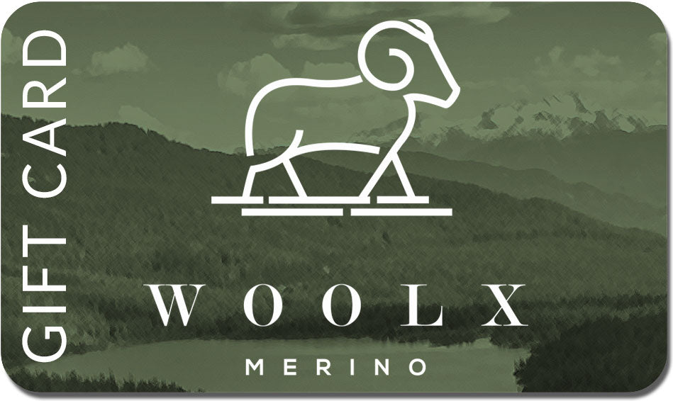 The Woolx Gift Card showcases a minimalist design of a ram on a hill with "WOOLX" and "MERINO" beneath, all set against a scenic mountain backdrop. The card is primarily green and adorned with clouds.