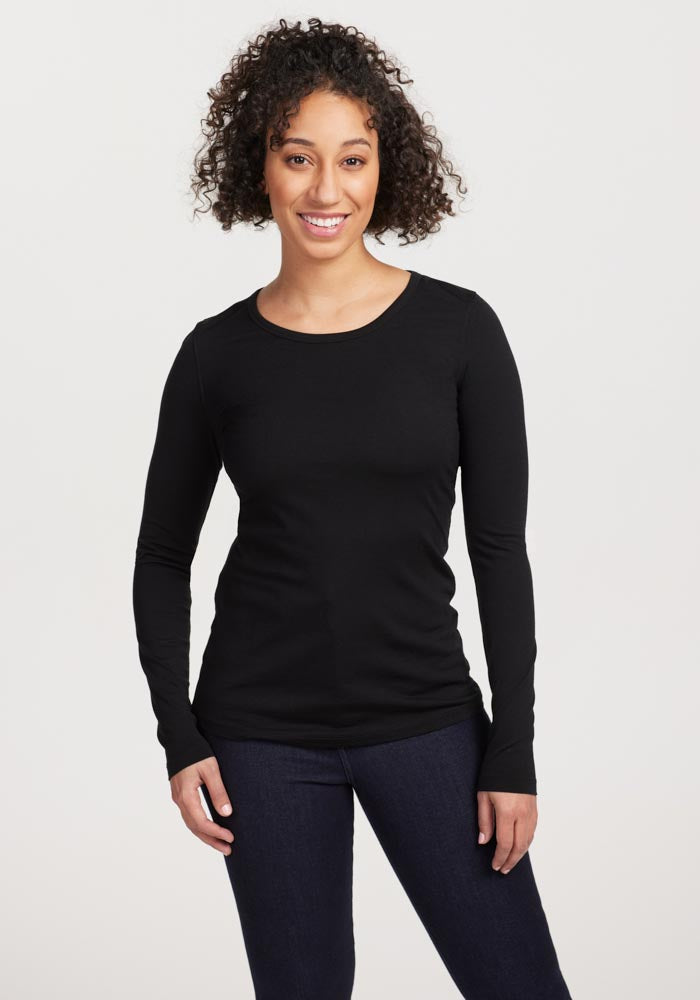 A person with curly hair is smiling and posing in a Woolx Remi Long Sleeve T-Shirt OLD in black, paired with dark pants against a plain backdrop. | Tori is 5'7", wearing a size S